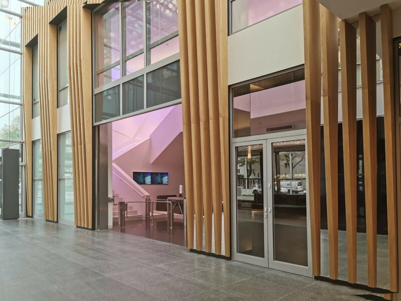 Innenansicht einer modernen Büro-Lobby mit vertikalen Holzlamellen, gläsernen Eingangstüren und einer Treppe mit rosa Beleuchtung.