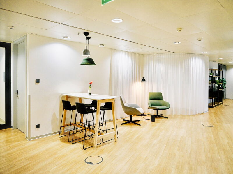 Heller und moderner Büro-Lounge-Bereich mit Stehtisch und Barhockern, bequemen grünen und grauen Sesseln, stilvollen Pendelleuchten und weißen Vorhängen im EuroPlaza.