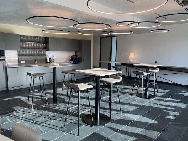 Heller, moderner Büro-Lounge-Bereich mit Stehtischen und Barhockern, einer Teeküche im Hintergrund und auffälligen runden Pendelleuchten.