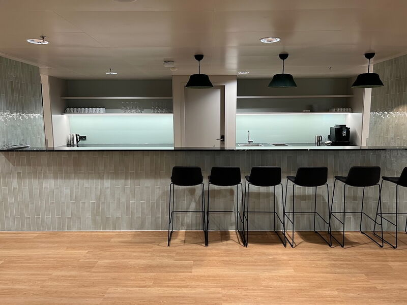 Moderne Büro-Teeküche mit langer Theke, schwarzen Barhockern, offenen Regalen und Kaffeemaschine vor hellgrünen Fliesenwänden.