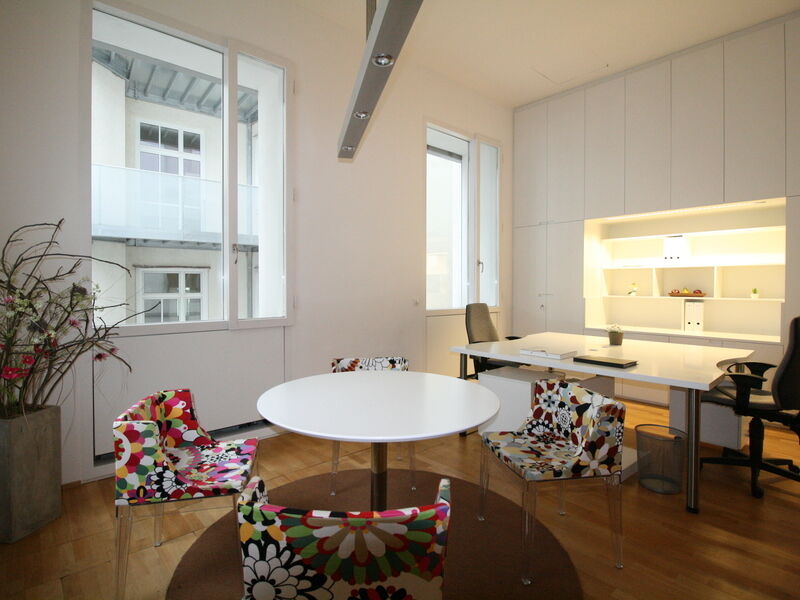 Ein helles, modernes Bürointerieur mit einem runden weißen Tisch, umgeben von bunten Blumenmustermöbeln, einem großen weißen Schreibtisch und Einbauschränken.