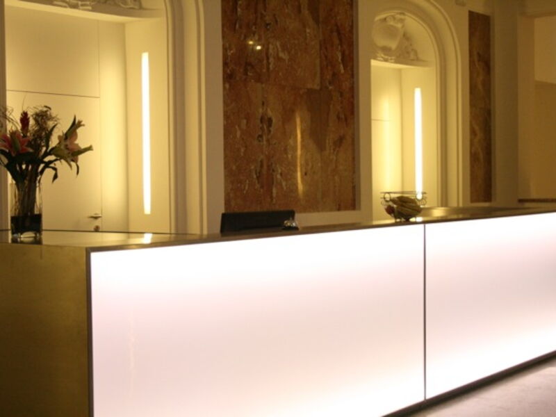 Stilvolle und moderne Hotelrezeption mit einer beleuchteten weißen Theke, Blumenvase und eleganten Marmorwandpaneelen im Hintergrund