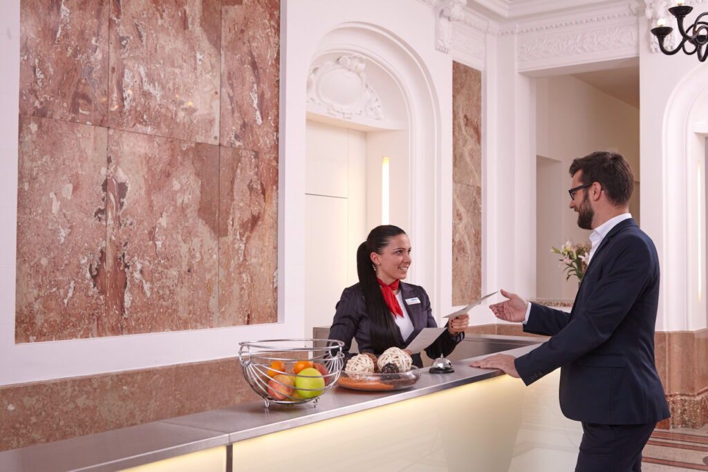 Friendly hotel receptionist handing documents to a smiling male guest at a modern reception desk in an elegant hotel lobby