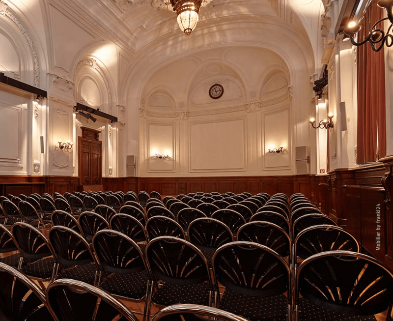 Spacious historic conference hall featuring ornate white arched ceilings, wood paneling, and rows of elegant chairs facing the front.