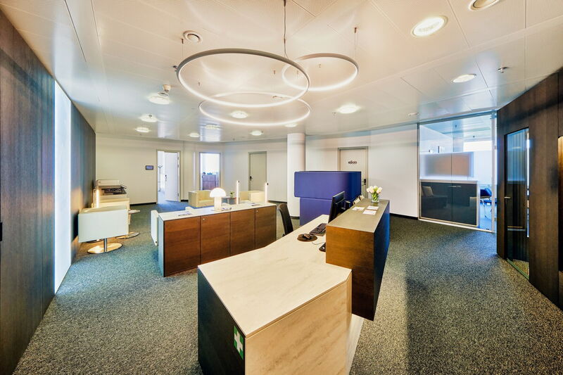 Geräumiger und moderner Büro-Empfangsbereich mit eleganten Holzschreibtischen, bequemen Sitzgelegenheiten und runden Designer-Deckenleuchten.
