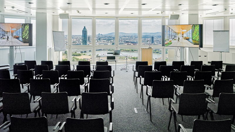 Heller, moderner Konferenzraum mit Stuhlreihen, großen Präsentationsbildschirmen und einem atemberaubenden Panoramablick auf die Wiener Skyline durch bodentiefe Fenster.