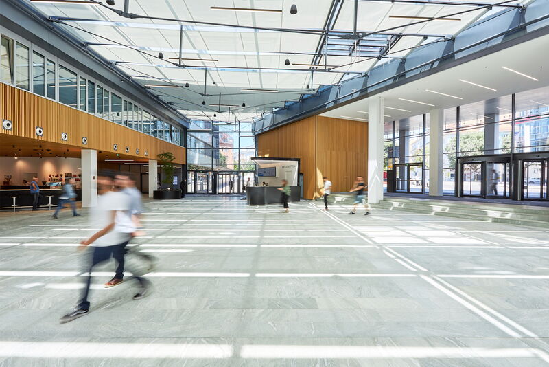 Helles und geräumiges modernes Büro-Atrium mit Innenbäumen, großem Glasdach, weißer Treppe und Personen auf dem Marmorboden.