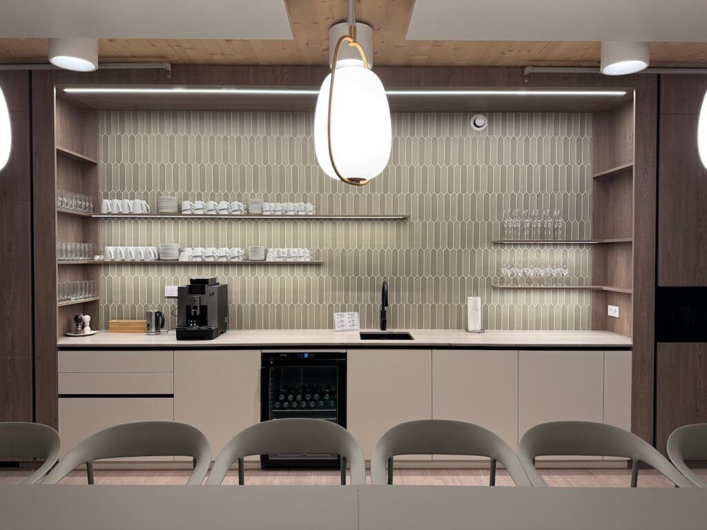 Stilvolle Büro-Teeküche mit hellgrüner Spiegelfliesen-Rückwand, Holzregalen, integriertem Weinkühlschrank, Kaffeemaschine und modernen Pendelleuchten.