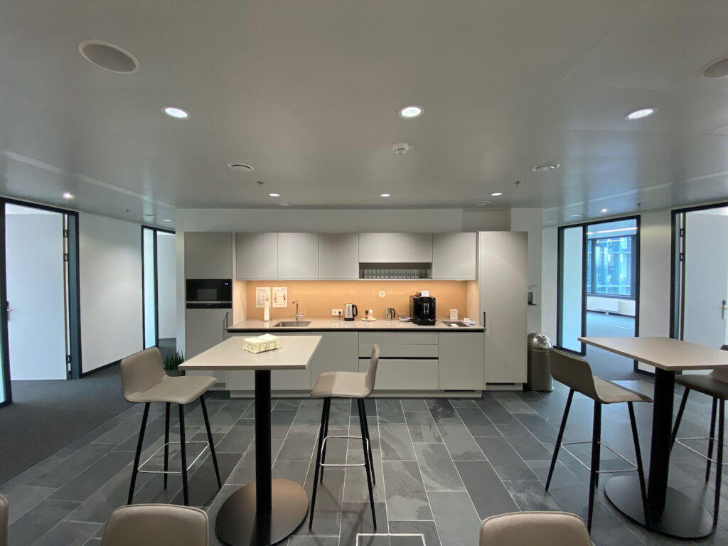 A modern, well-equipped office kitchenette with grey cabinetry, a built-in coffee machine, sink, and high-top seating tables with stools on a dark tile floor.