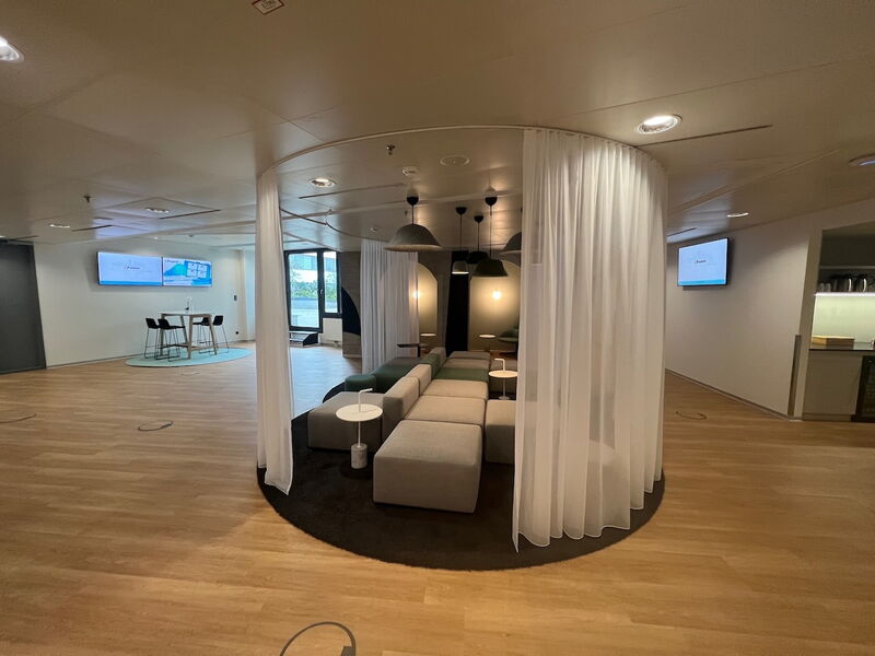 Geräumige moderne Büro-Lounge mit modularen, grau-grünen Sitzgelegenheiten, eingerahmt von halbdurchsichtigen weißen Vorhängen, Holzboden und Besprechungsbereichen im Hintergrund.