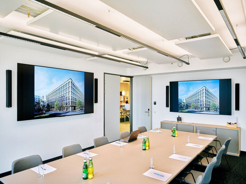 Moderner Konferenzraum mit einem großen Holztisch, ergonomischen Stühlen, Wasserflaschen und zwei großen Wandbildschirmen mit Architekturdarstellungen.