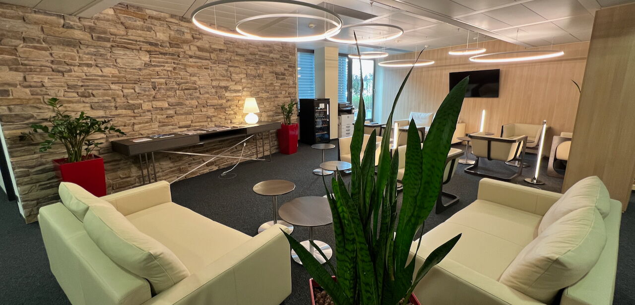 Spacious modern office lounge featuring comfortable white sofas, a rustic stone accent wall, stylish lighting, and indoor plants.
